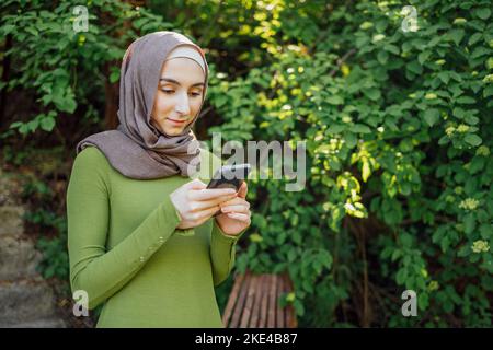 Jeune étudiante musulmane moderne portant le hijab dans le parc, tenant un téléphone dans la main et souriant Banque D'Images