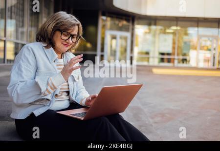 Bonne femme d'âge moyen effectuant des appels vidéo sur ordinateur portable, en agitant à l'écran, en discutant en ligne avec des personnes qui la distance. Femme âgée souriante aux cheveux gris wav Banque D'Images