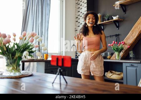 Une femme vlogger influenceur noir s'assoit à la maison en regardant la caméra, une jeune femme blogger accueille les fans et parle à la cuisine et en regardant le smar Banque D'Images