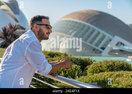 Homme élégant avec chemise blanche et lunettes de soleil regardant loin se pencher sur un balustrade avec la ville des arts et des sciences de valence en arrière-plan. Banque D'Images