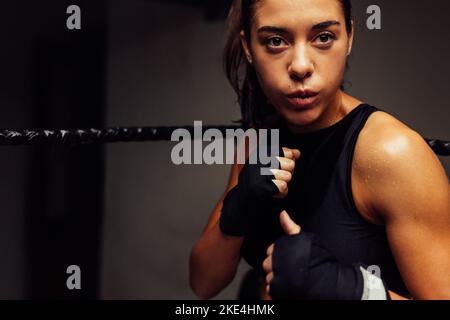 Femme sportive debout dans un anneau de boxe avec les mains enveloppées avant un combat. Boxeur féminin ayant une séance d'entraînement dans un gymnase de boxe. Banque D'Images