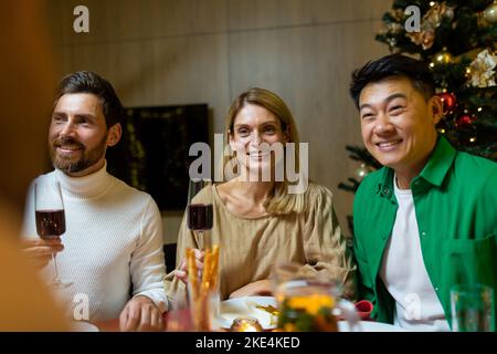 De jeunes gens interraciaux heureux assis à la table du nouvel an festif. Ils tiennent des verres de vin, parlent, sourient, célèbrent Noël avec des amis. Banque D'Images