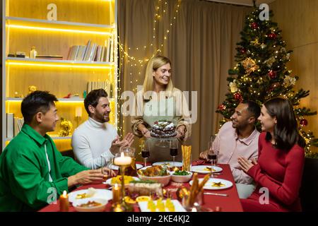 Une jeune femme, la propriétaire de la maison, traite les invités, tient un gâteau dans ses mains, le met sur la table. Des hommes et des femmes s'assoient à la table du nouvel an, applaudissent la femme, sourient. Banque D'Images