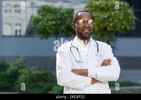 Portrait d'un médecin expérimenté mature à l'extérieur d'une clinique moderne, un homme afro-américain portant des lunettes et un manteau médical regarde l'appareil photo et souriant amical avec ses bras croisés. Banque D'Images