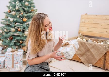 Élégante et moderne, cette femme millénaire est assise à l'arbre de Noël artificiel, tient un cadeau entre ses mains, sourit. Rouge à lèvres sur les lèvres. Ambiance festive et écologique Banque D'Images