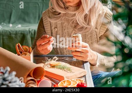 Emballage des cadeaux de noël. Femme blonde emballage des cadeaux en carton recyclé et décoré avec des oranges séchées et des branches de sapin près de l'arbre de Noël. Banque D'Images