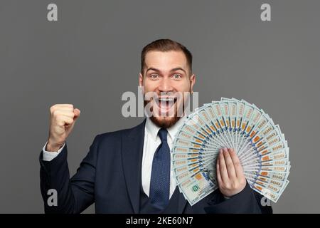 Beau homme d'affaires heureux en costume avec un fan de dollars US l'argent criant sur fond gris studio. Concept de gain, de plaisir et de réussite Banque D'Images