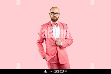 Chauve homme en costume rose et lunettes posant avec la main dans la poche sur fond rose studio Banque D'Images