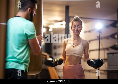 Mettre des gants de boxe en forme de fille en souriant. Instructeur de fitness masculin avec une cliente pratiquant kickbox avec sac à puncing dans un studio de fitness. Banque D'Images