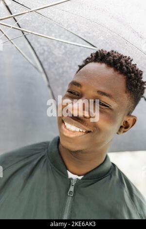 Jeune Africain américain avec parapluie, sous la pluie, souriant dans la rue à l'extérieur. Automne ou printemps Banque D'Images