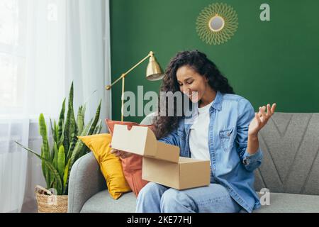 Jeune belle femme hispanique à poil dur a reçu le paquet de la boutique en ligne, heureux et satisfait de la livraison assis sur le canapé et heureux dans le salon avec mur vert. Banque D'Images