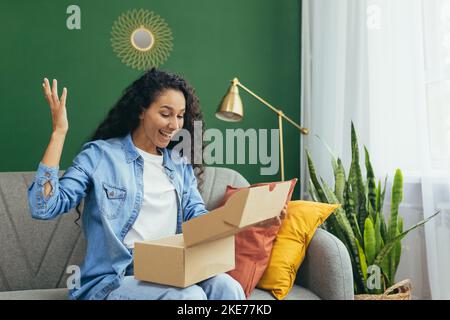 Jeune belle femme hispanique à poil dur a reçu le paquet de la boutique en ligne, heureux et satisfait de la livraison assis sur le canapé et heureux dans le salon avec mur vert. Banque D'Images