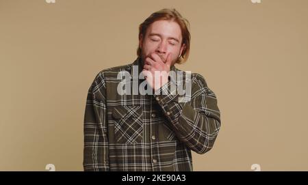 Fatigué beau barbu homme dans la chemise à carreaux bâilling endormi inattentif sentiment somnolent paresseux ennuyé gaping souffrant du manque de sommeil. Jeune garçon blond isolé sur fond beige studio Banque D'Images