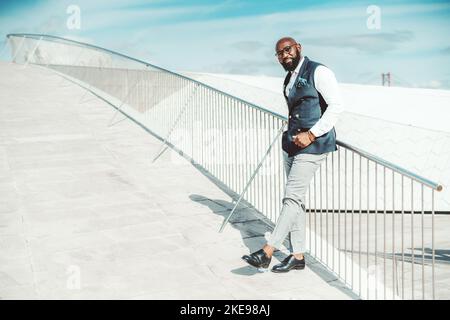 Un homme d'affaires afro-américain à la mode et à la barbe chauve-américaine dans un élégant costume habillé ajusté avec un gilet est penché sur la clôture sur le stre Banque D'Images