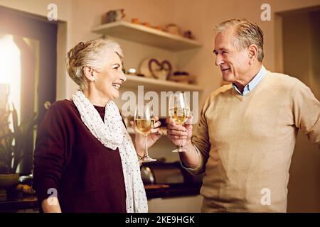 Heres à une vie passée ensemble. Un couple âgé bavardant et buvant du vin ensemble tout en se tenant dans leur cuisine. Banque D'Images