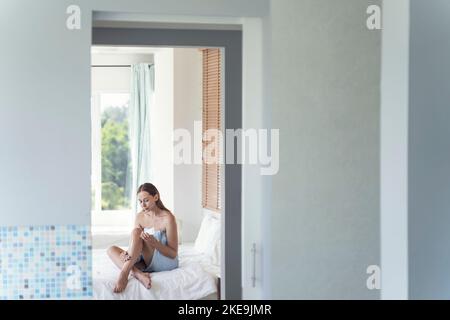 Une jeune femme applique de la crème sur ses jambes tout en étant assise sur le lit. Une jeune femme prend soin du corps tout en étant assise sur un lit dans une pièce lumineuse. Soins personnels. Banque D'Images