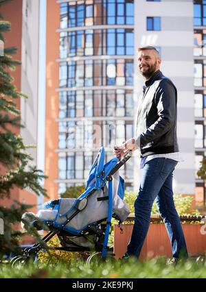 Le père passe du temps avec un nouveau-né à l'extérieur. Jeune homme barbu marchant avec bébé poussette dans la rue dans la cour moderne des immeubles résidentiels de la ville. Banque D'Images