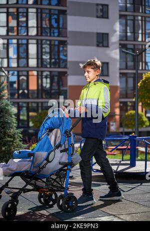 Jeune garçon soucieux de passer du temps avec la sœur du nouveau-né à l'aire de jeux. Un adolescent marchant avec bébé dans une poussette dans la rue dans la cour moderne des immeubles résidentiels de la ville. Banque D'Images