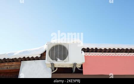 Inverseur recouvert de neige unité extérieure de climatisation, vue rapprochée sur le toit. Un toit enneigé d'une maison en bois sur fond bleu ciel Banque D'Images