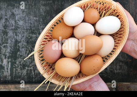 Les mains des agriculteurs tiennent les oeufs de poules domestiques dans le panier sur fond de texture en bois ancienne. Œufs blancs et bruns rouges naturels. Gros plan, vue de dessus. Banque D'Images