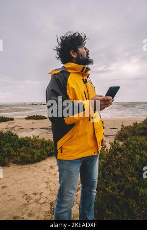 Un homme debout dans le vent dans les loisirs en plein air seul en utilisant le téléphone mobile et la connexion Internet d'itinérance. Veste jaune. Style de vie sauvage. Wint Banque D'Images