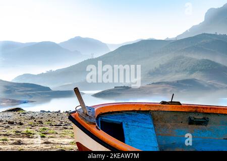 Vieux canot en bois, bateau bleu, orange et blanc coincé sur le rivage de terre sèche dans le barrage du lac de Bouzegza Keddara avec des montagnes brumeuses reflétées. Banque D'Images