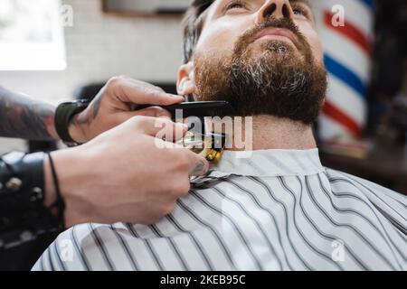 homme barbu près de coiffeur se coupant le cou avec une tondeuse à cheveux dans barbershop, image de stock Banque D'Images