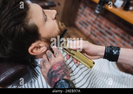 vue en grand angle du coiffeur tatoué taille de la barbe de l'homme avec tondeuse à cheveux, image de stock Banque D'Images