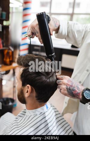 coiffeur tatoué dans montre-bracelet sèche cheveux de client dans barbershop, image de stock Banque D'Images