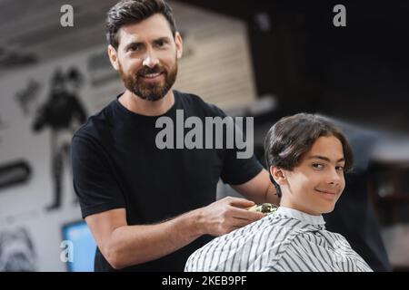 Coiffeur souriant tenant la tondeuse près du client et regardant l'appareil photo dans le salon de beauté, image de stock Banque D'Images