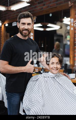 coiffeur positif montrant comme le geste et regardant la caméra près de l'adolescent dans le cap de coiffure, image de stock Banque D'Images