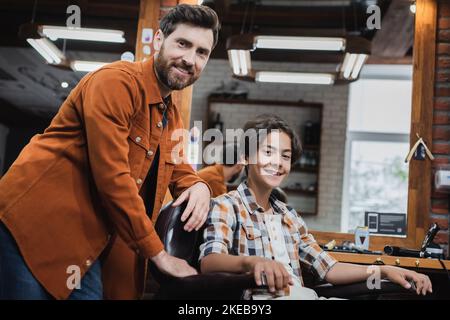 Client ado souriant et coiffeur regardant la caméra dans le barbershop, image de stock Banque D'Images