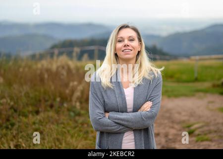 Belle femme blonde d'âge moyen, douce et souriante, aux bras croisés se trouve sur une montagne dans la Forêt Noire Banque D'Images