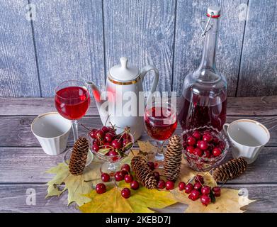 Sur une table en bois, il y a une bouteille et des verres remplis de vin rouge, une cafetière et des tasses, un vase avec des cerises mûres, des feuilles d'érable jaune et des cônes de sapin. Banque D'Images