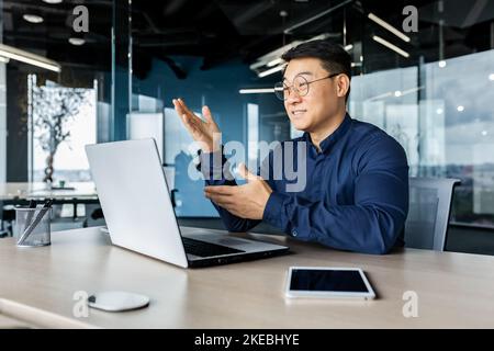 Homme d'affaires asiatique prospère qui parle sur appel vidéo à l'aide d'un ordinateur portable pour communiquer à distance avec ses collègues, patron en chemise souriant et gestimant avec les mains, réunion en ligne avec ses partenaires. Banque D'Images