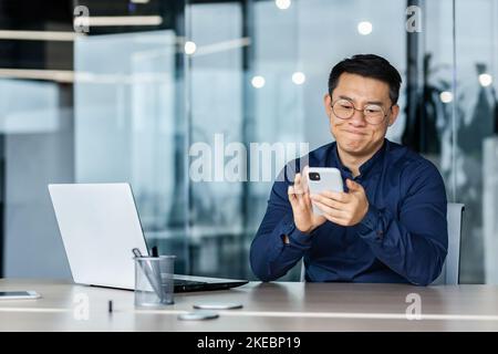 Un homme d'affaires asiatique insatisfait a eu de mauvaises nouvelles, un homme lisant sur un smartphone assis dans un bureau moderne avec un ordinateur portable, un patron déçu par le téléphone contrarié. Banque D'Images