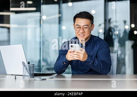 Homme d'affaires asiatique prospère travaillant dans un immeuble de bureaux moderne, homme souriant et heureux en utilisant un smartphone pour parcourir des pages en ligne, homme dans une chemise et des lunettes avec ordinateur portable. Banque D'Images