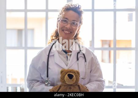 Portriat de jeune femme médecin en uniforme et stéthoscope tenant l'ours en peluche contre la fenêtre à la clinique. Professionnel de la santé ou professionnel de la santé confiant dans les lunettes à l'hôpital Banque D'Images