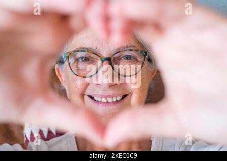 Gros plan d'une femme âgée faisant un geste de forme de coeur avec les mains. Femme âgée heureuse à la retraite qui fait le symbole de la forme du cœur avec ses deux mains et regardant l'appareil photo Banque D'Images