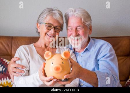 Gros plan d'un couple senior montrant une banque de pigeons pour économiser de l'argent. Couple de personnes âgées tenant une banque de piggy pour l'investissement et le concept de planification future. Heureux couple à la retraite tenant ensemble une tirelire à la maison Banque D'Images