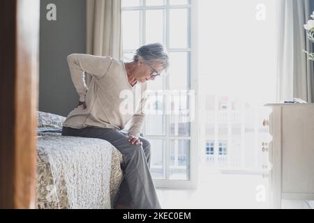 Femme âgée souffrant de mal de dos après le sommeil, frottement de muscles raides, ancienne femme assise sur le lit touchant le bas du dos sensation d'inconfort en raison du lit inconfortable à la maison Banque D'Images