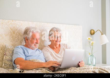 Un couple senior souriant navigue et regarde du contenu sur les réseaux sociaux à l'aide d'un ordinateur portable au lit à la maison. Un vieux mari et une femme détendus passent du temps au lit, partageant un film en ligne dans la chambre à coucher dans un appartement moderne Banque D'Images