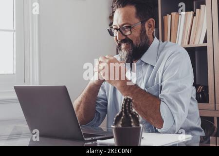 Beau homme adulte barbu souriant sur la réception ou la lecture de bonnes nouvelles à l'aide d'un ordinateur portable au bureau à domicile. Un homme d'affaires caucasien heureux travaillant sur un ordinateur portable vérifiant les e-mails assis à la réception au bureau Banque D'Images