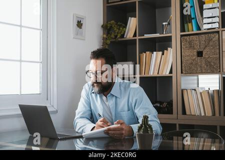 Joyeux jeune homme d'affaires caucasien souriant travaillant en ligne regardant le podcast du webinaire sur ordinateur portable et apprentissage formation cours conférence téléphonique prendre des notes assis au bureau, concept d'apprentissage en ligne Banque D'Images