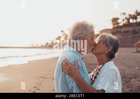 Gros plan de deux personnes âgées mignonnes et heureuses qui s'amusent et s'amusent ensemble un jour de coucher de soleil à la plage. Couple adulte amoureux embrassant avec le coucher de soleil à l'arrière-plan. Banque D'Images