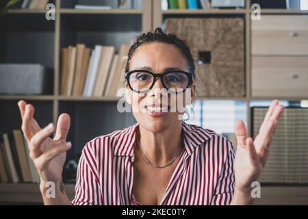 Portrait de la tête une femme d'affaires confiante porte des lunettes regardant l'appareil photo et parlant, mentor intervenant tenant une leçon en ligne, expliquant, assis à un bureau en bois dans un cabinet moderne Banque D'Images