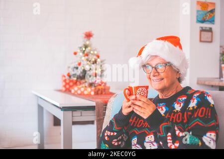Portrait d'une femme âgée à la retraite souriante en chapeau de père noël et vêtements chauds célébrant noël à la maison. Une vieille femme qui souriait en regardant l'appareil photo. Femme âgée appréciant les vacances de noël Banque D'Images