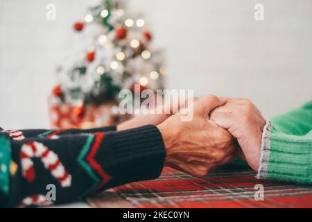 Couple senior en vêtements chauds tenant les mains les unes contre les autres devant l'arbre de noël décoré à la maison. Un vieux couple hétérosexuel romantique aimant célébrant ensemble le festival de noël Banque D'Images