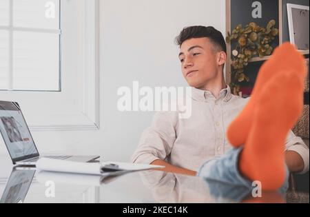 Un adolescent satisfait est heureux de terminer son travail avec un ordinateur portable à la maison, lève les mains et met les pieds sur la table, se détend après une dure journée de travail en prévision d'un congé de fin de semaine, d'une journée de travail détendue, sans stress Banque D'Images