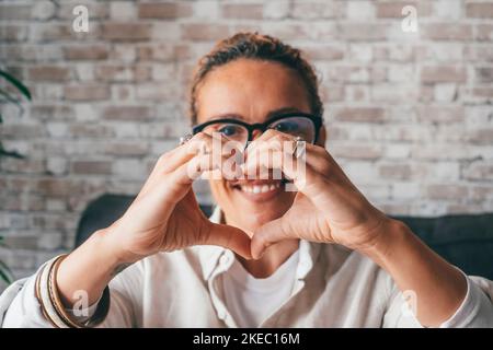 Bonne jeune femme romantique regardant la caméra à travers la forme de coeur fait de doigts joints. Portrait de la femme caucasienne montrant son amour et son affection en faisant signe d'amour avec les mains Banque D'Images